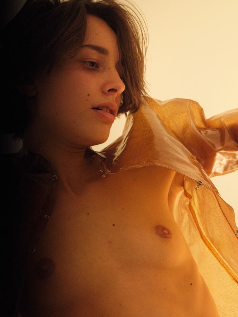 Low-angle, medium shot of a young adult with short brown hair wearing a sheer brown top against a light background in a photo studio.