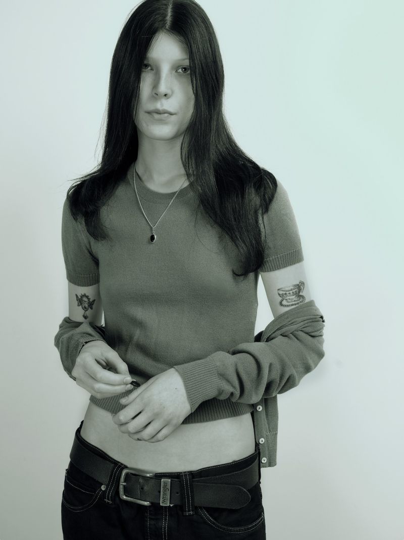 A young woman with long dark hair wearing a green top, black belt, and silver necklace against a plain white background.