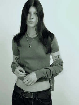 A young woman with long dark hair wearing a green top, black belt, and silver necklace against a plain white background.