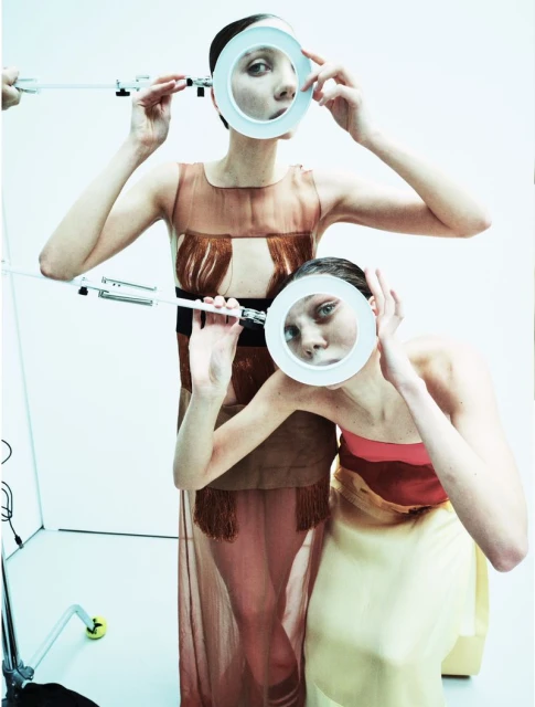 Two fashion models pose in a photo studio, holding white circular props near their faces, wearing sheer dresses, against a plain white background.