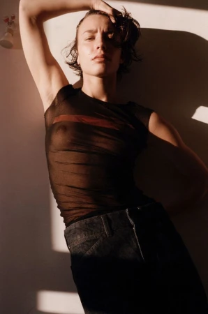 Model wearing a sheer black top and dark trousers posing against a sunlit wall in an editorial fashion photograph.