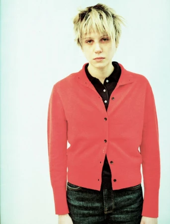 Model with blonde hair wearing a red button-down cardigan and dark jeans against a plain light background