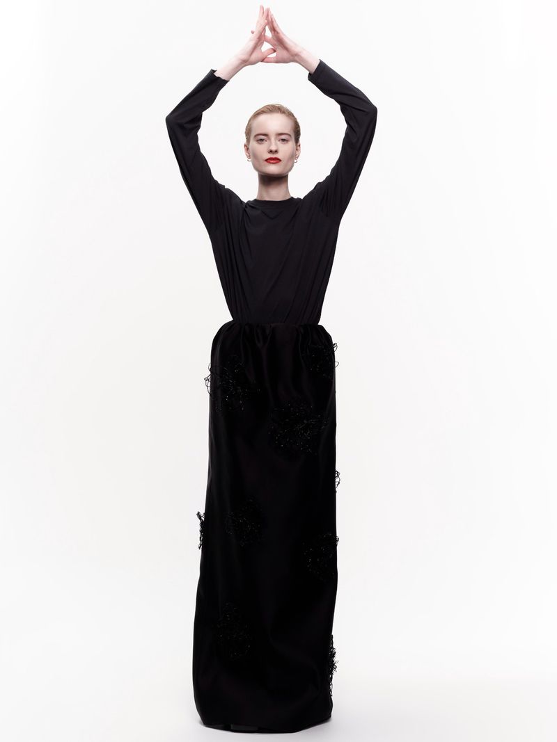 A woman stands in a black gown against a white background, hands above her head forming a triangular shape in a fashion pose.