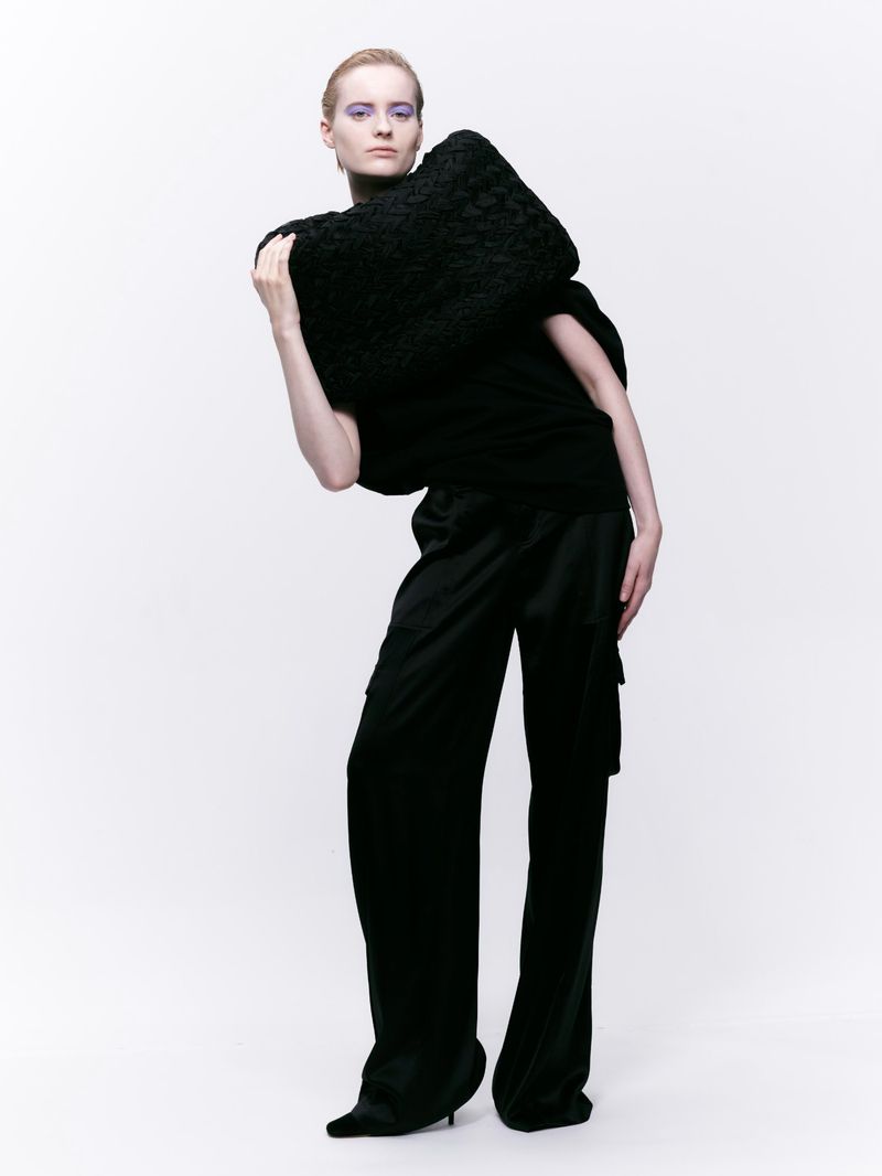 Fashion model posing in a studio wearing a chunky black sweater, wide-leg black pants, and black heeled shoes against a white backdrop.