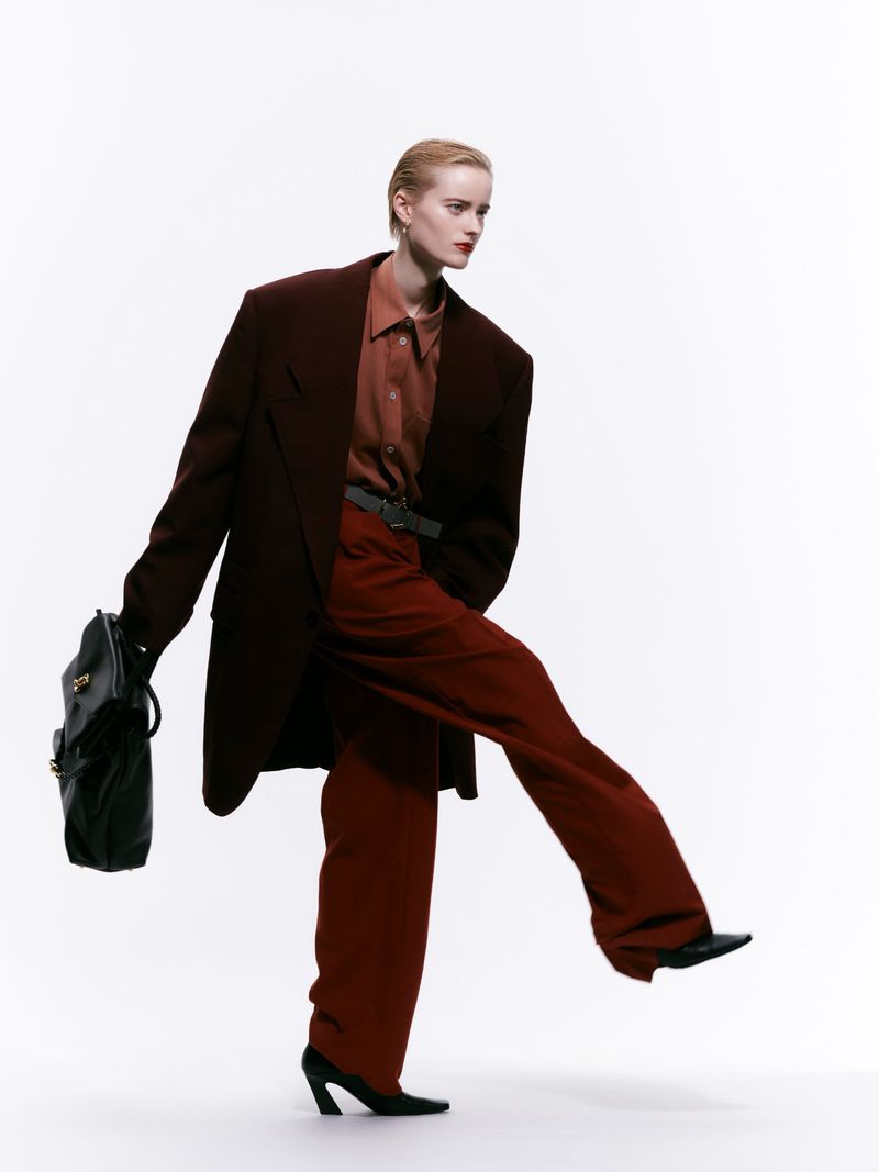 A person in a brown jacket and pants, posing against a white background, holding a black purse with a confident, dynamic stance.