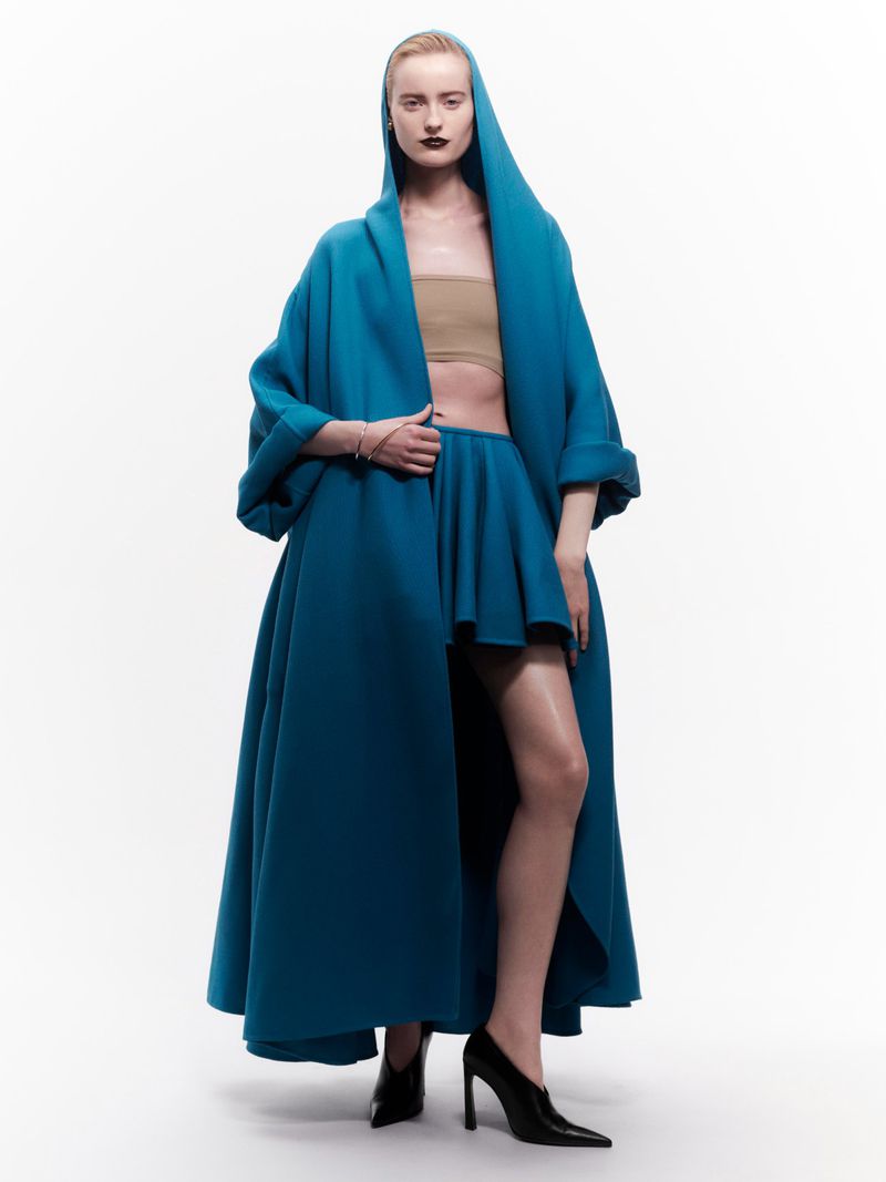 Fashion shot of a woman in a blue hooded cape, strapless top, and matching skirt against a white background in a studio setting.