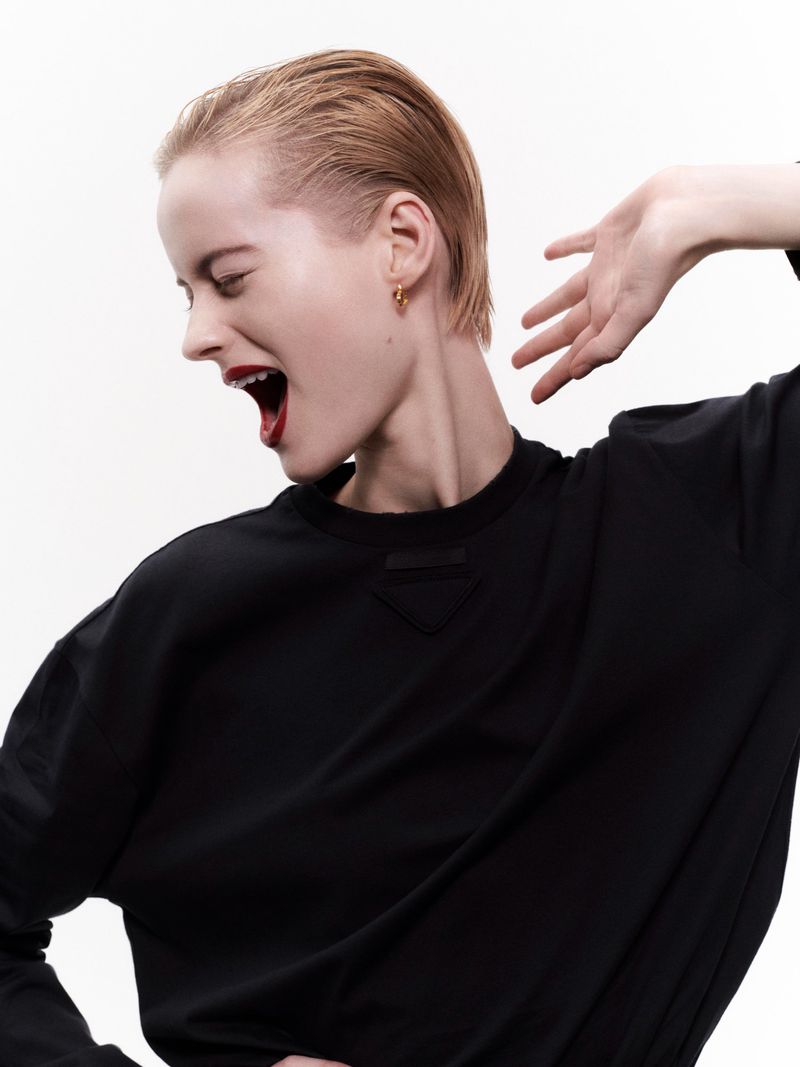 Woman in profile with slicked-back hair, gold hoop earring, black top, and red lipstick against a solid white studio backdrop.