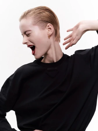 Woman in profile with slicked-back hair, gold hoop earring, black top, and red lipstick against a solid white studio backdrop.