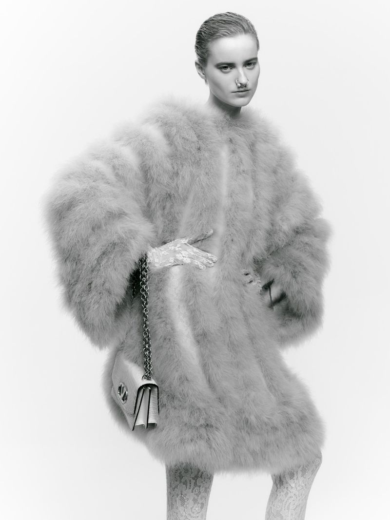 Fashion shot of a model wearing a furry coat and holding a purse against a white background in a studio environment.