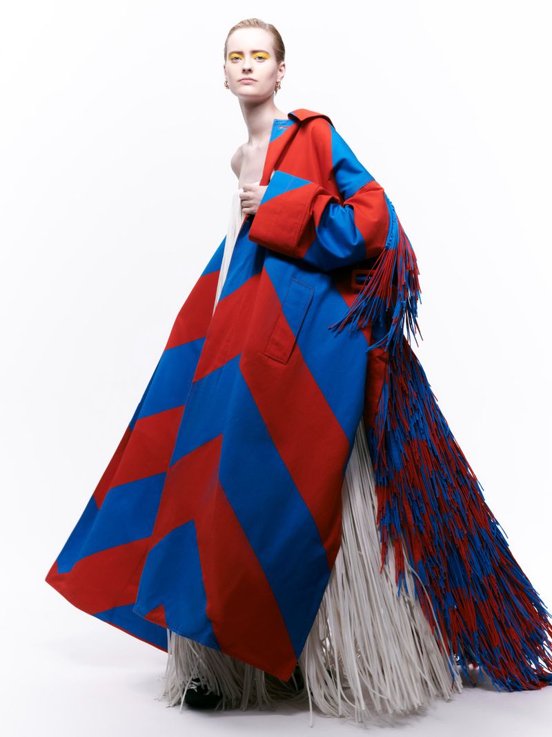 Fashion model poses in a vibrant blue and red geometric patterned coat with long fringe against a clean white backdrop in studio lighting.