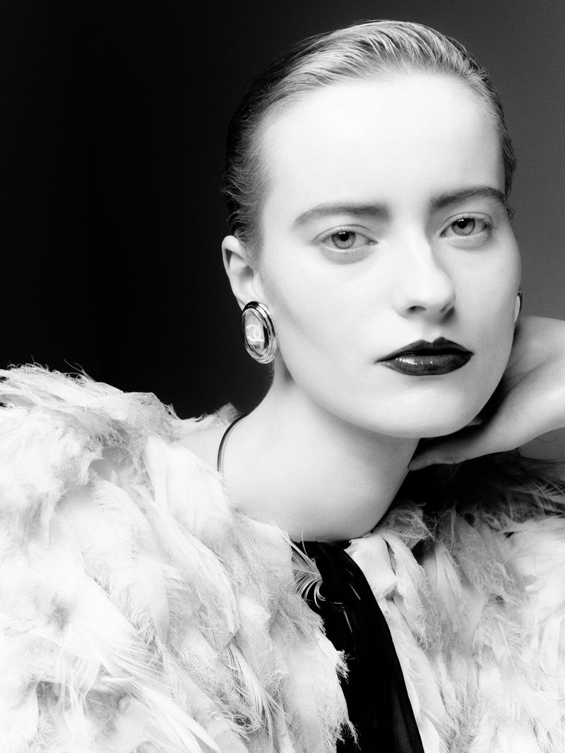 Black and white portrait of a female model with pale skin, dark lipstick, and a feathered jacket posing in front of a dark background.