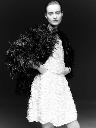 Model in a white textured dress and dark fringed jacket against a dark background for Puss Puss Magazine