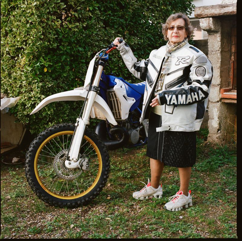 Elderly woman poses with a white and blue Yamaha dirt bike outdoors, wearing a riding jacket, glasses, and sneakers in daylight.