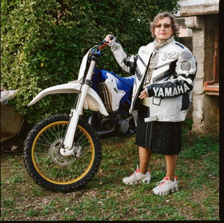 Elderly woman poses with a white and blue Yamaha dirt bike outdoors, wearing a riding jacket, glasses, and sneakers in daylight.