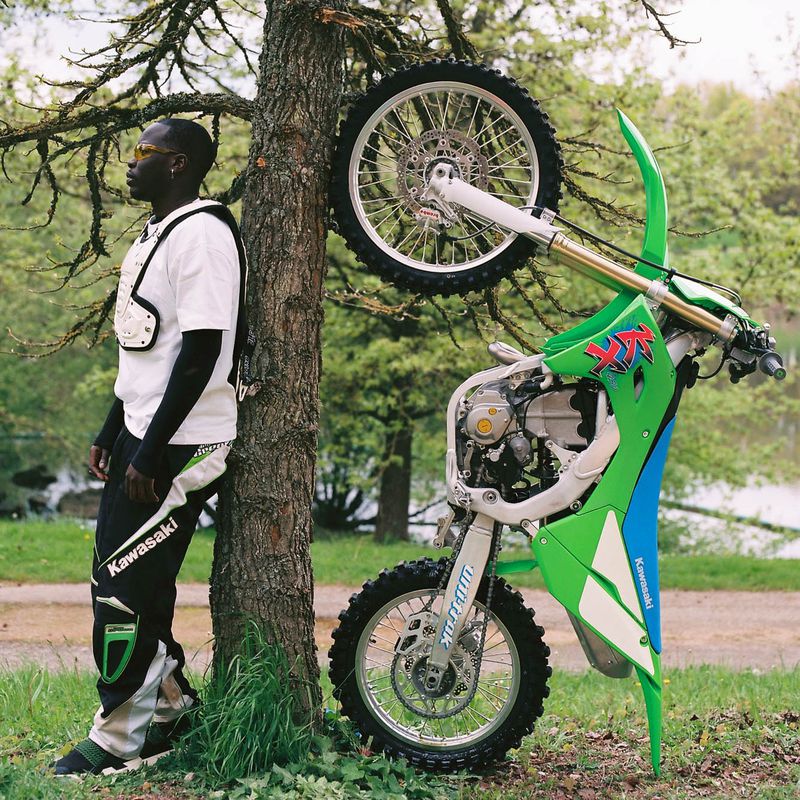 Man in Kawasaki racing pants stands by a tree supporting an upside-down Kawasaki dirt bike with a green and white color scheme in an outdoor setting.