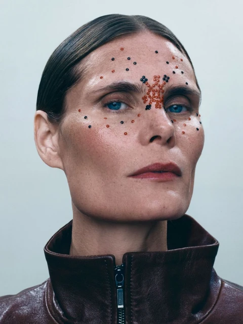 Close-up portrait of a fair-skinned adult woman with slicked-back brunette hair and artistic beaded makeup; wearing a brown leather jacket.