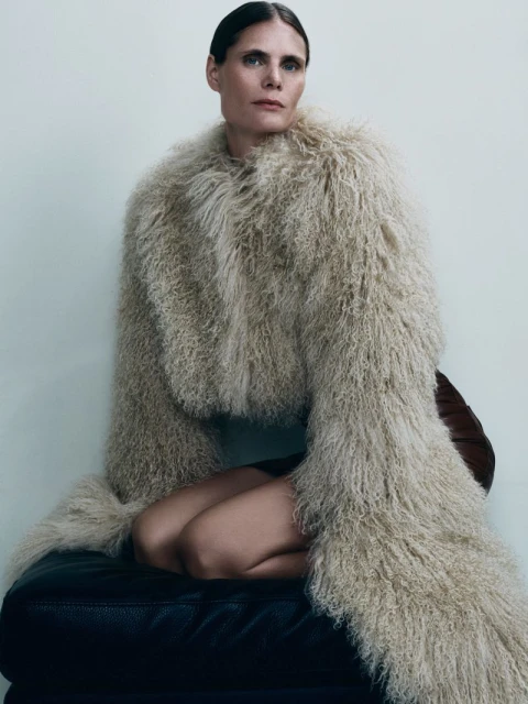 Woman in a fur coat sits on a black leather ottoman with her legs crossed. Her hands rest on her legs, and she gazes off into the distance.