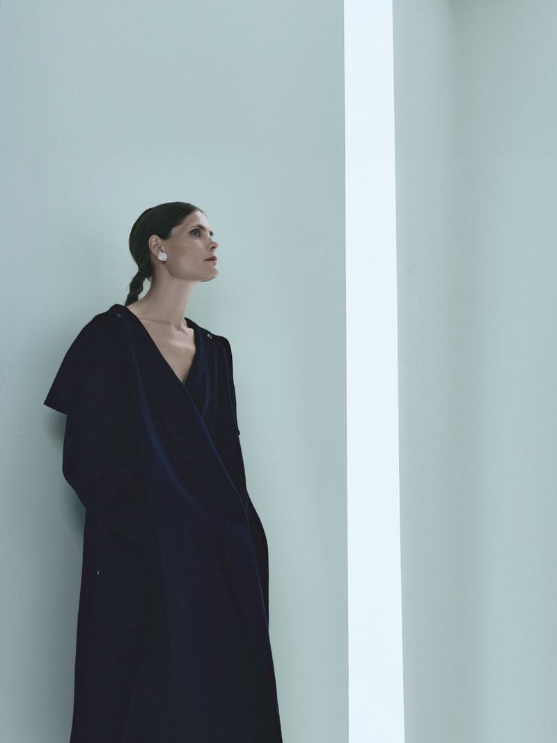 Elegant adult woman in a dark robe stands near a white wall with light reflection, looking up with a neutral expression in an indoor setting.