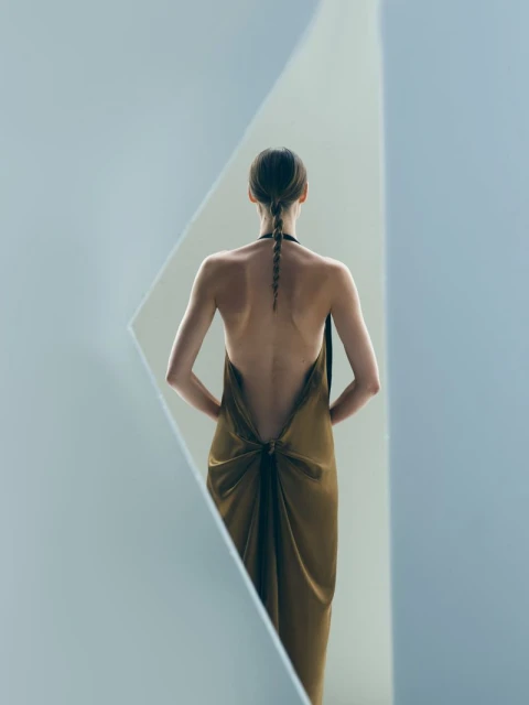 Back view of a slender adult woman in an olive dress with braided hair, framed by converging geometric shapes and cool lighting.