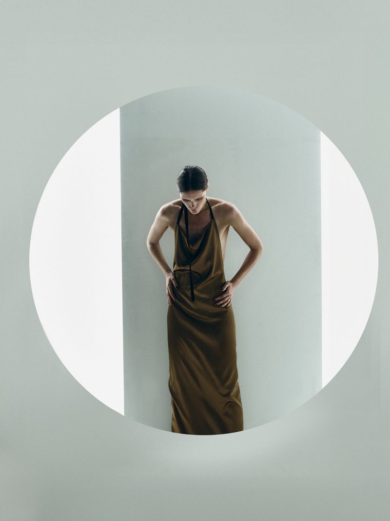 An adult woman with dark hair stands indoors wearing a long sleeveless earth-tone dress, hands on hips, in a bright and minimalist setting.