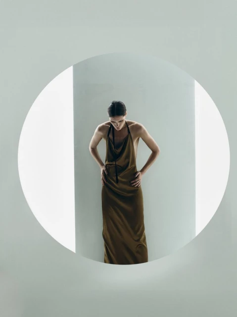 An adult woman with dark hair stands indoors wearing a long sleeveless earth-tone dress, hands on hips, in a bright and minimalist setting.