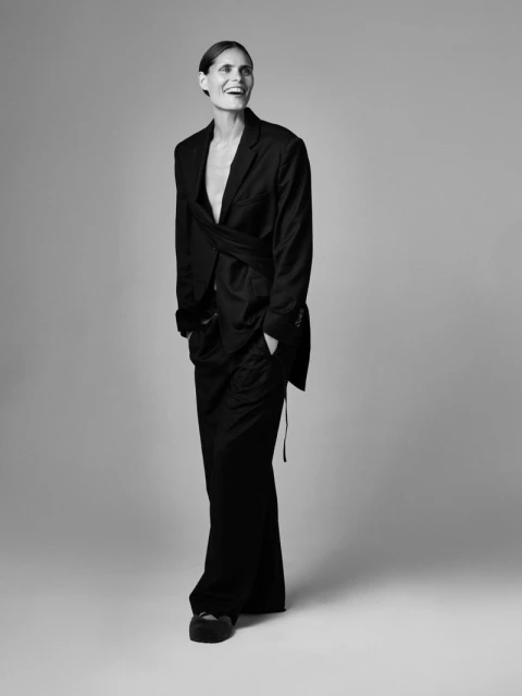 Monochrome fashion portrait of an adult woman in an oversized blazer and wide-leg trousers, striking a relaxed pose in a studio setting.
