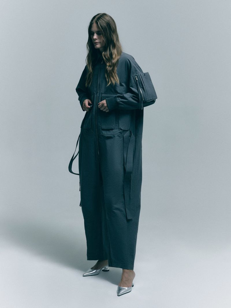 Woman in a dark grey utility jumpsuit with multiple pockets and silver heels against a neutral grey background in a studio setting.
