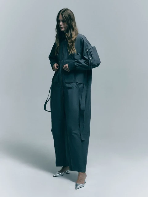 Woman in a dark grey utility jumpsuit with multiple pockets and silver heels against a neutral grey background in a studio setting.