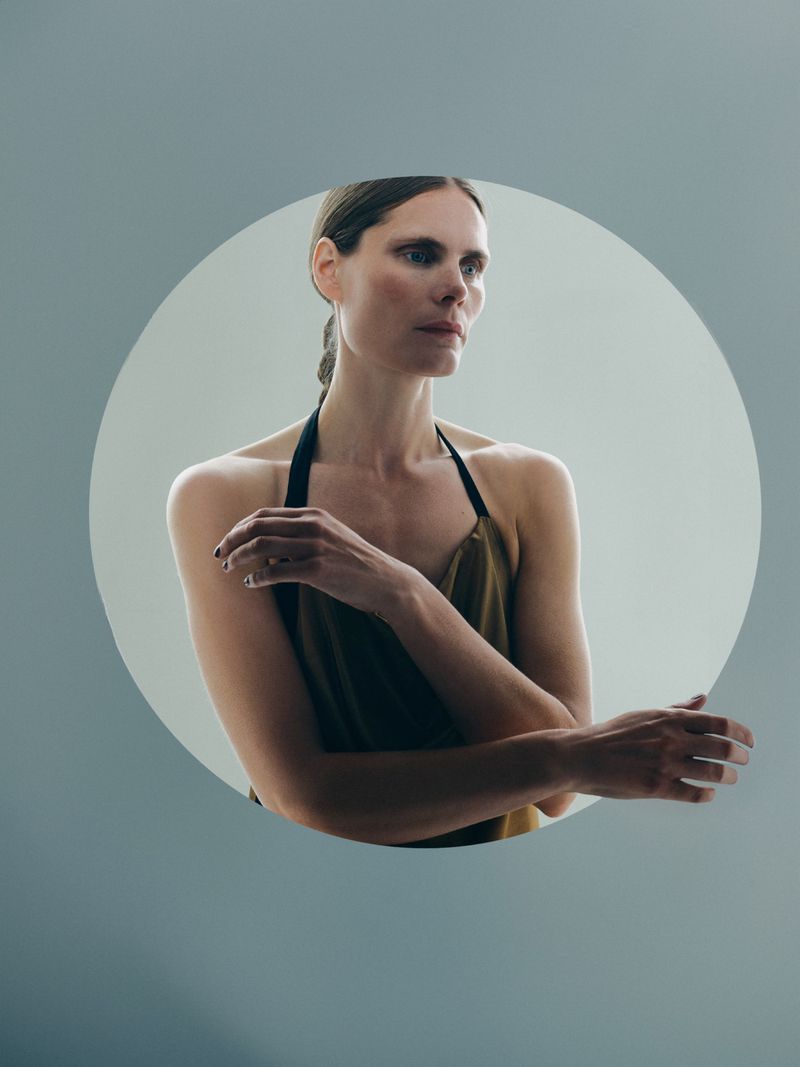 Woman in olive green top with arms crossed in a portrait against a light grey backdrop, showcasing soft studio lighting and minimalist style.