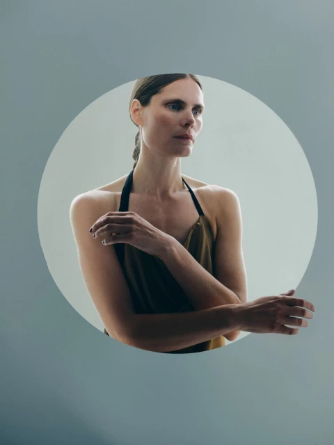 Woman in olive green top with arms crossed in a portrait against a light grey backdrop, showcasing soft studio lighting and minimalist style.