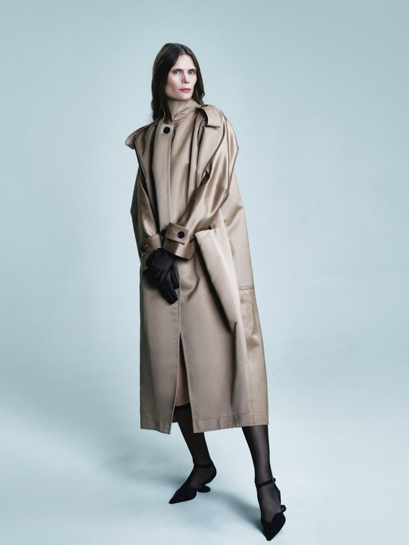 An adult female model poses in a tan trench coat, black gloves, black heels, and sheer black hosiery against a gradient blue backdrop.
