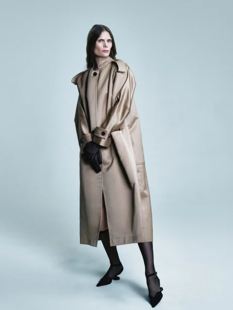 An adult female model poses in a tan trench coat, black gloves, black heels, and sheer black hosiery against a gradient blue backdrop.