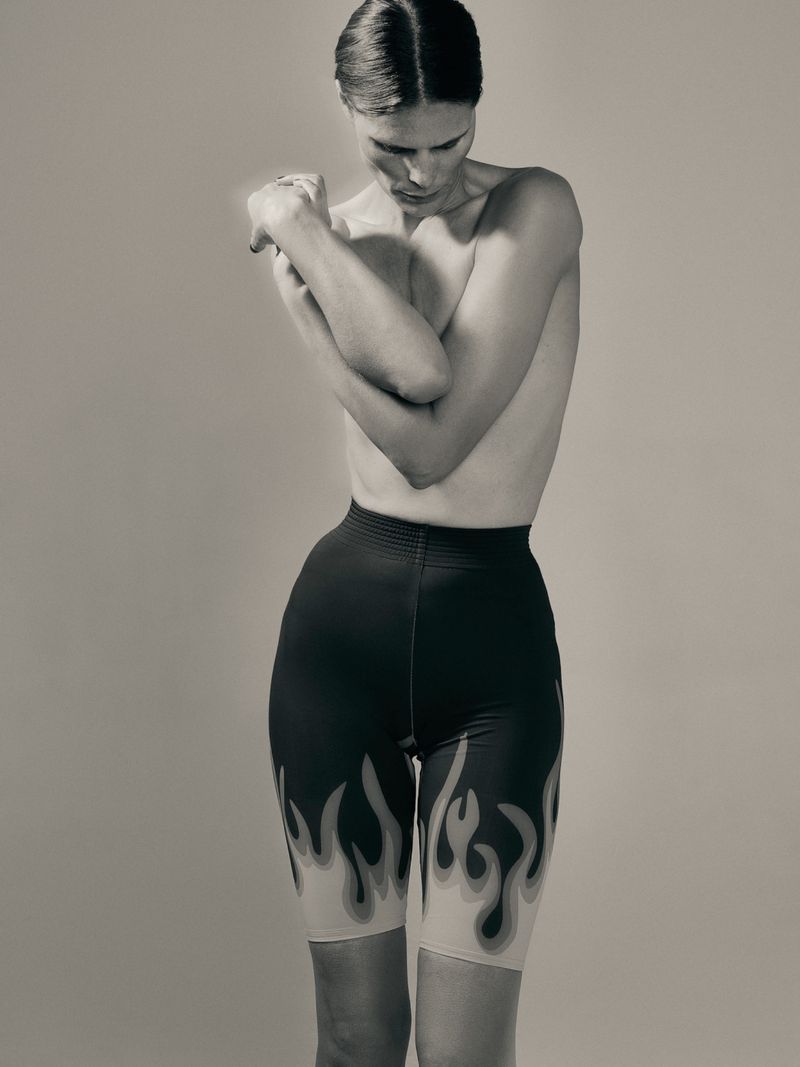 A slender adult female poses in a photo studio wearing flame-printed black shorts, captured in a medium shot with neutral background.