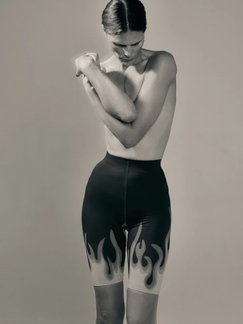A slender adult female poses in a photo studio wearing flame-printed black shorts, captured in a medium shot with neutral background.