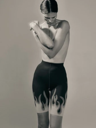 A slender adult female poses in a photo studio wearing flame-printed black shorts, captured in a medium shot with neutral background.