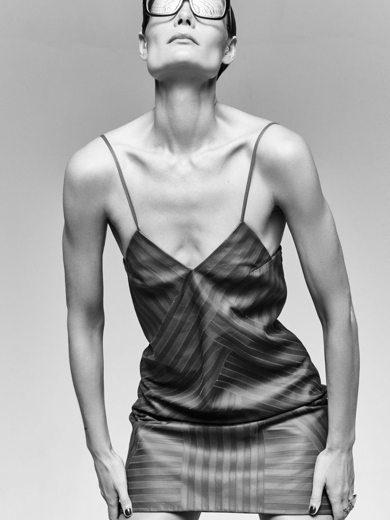 Black and white portrait of a slim, adult female model in a striped mini dress and large glasses, looking up with neutral expression.