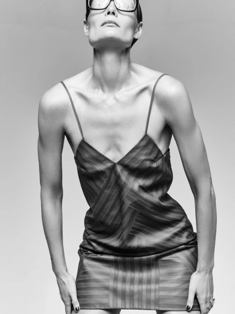Black and white portrait of a slim, adult female model in a striped mini dress and large glasses, looking up with neutral expression.