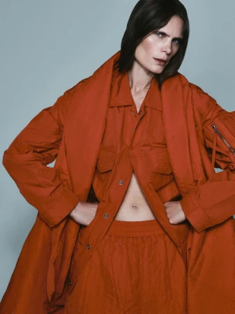 Model Adina Fohlin in a rust orange utility coat and matching trousers against a pale grey seamless background.
