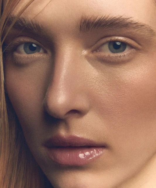 Close-up portrait of a young adult woman with light skin and blue eyes, soft pink lips, and blonde hair, evenly lit in a studio setting.