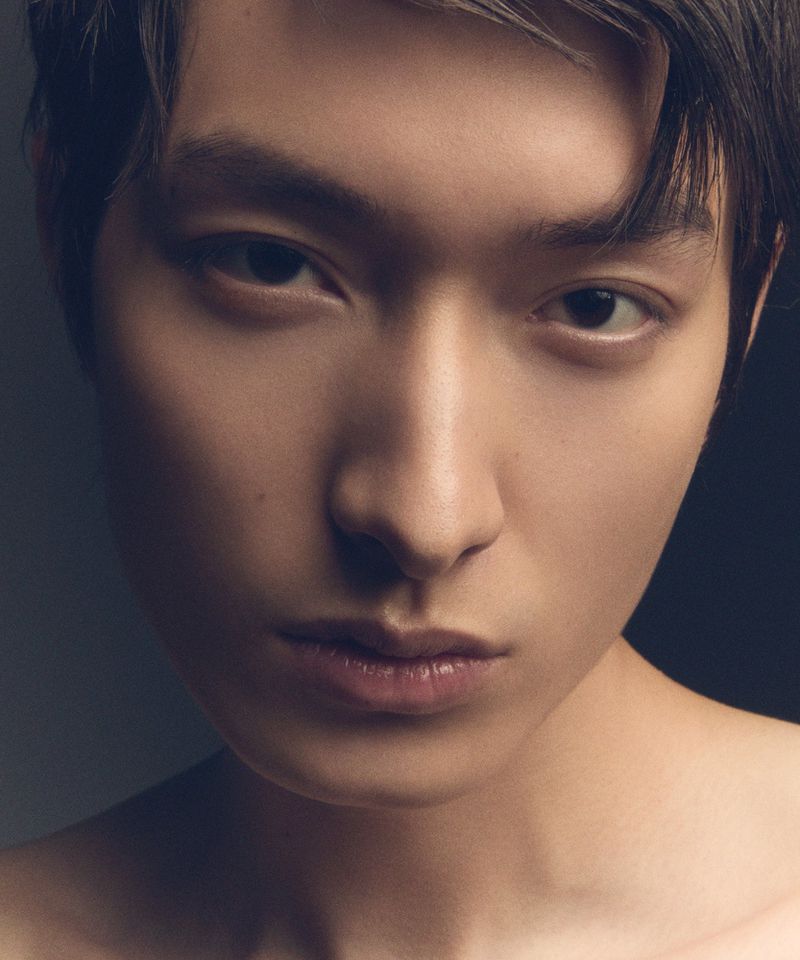 Close-up portrait of a young adult male with dark hair and light skin, looking directly at the camera with a neutral expression, dark background.