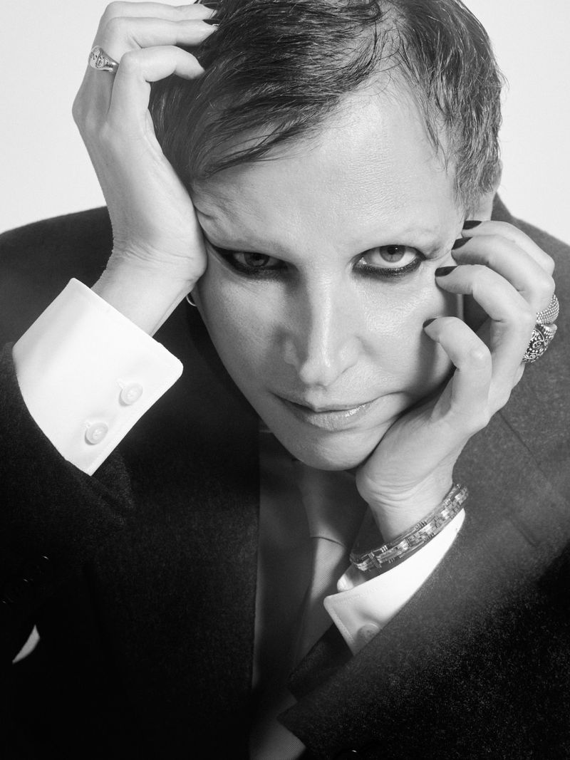 Monochrome portrait of an adult with hands framing the face, wearing a dark coat and rings, eyeliner makeup, against a neutral background.