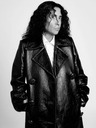 Portrait of a woman with dark curly hair wearing a shiny black coat and white shirt against a white background