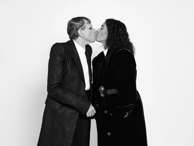 Black and white portrait of two adults in long coats kissing against a white backdrop, embodying high fashion and elegant style.