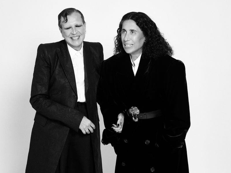 Two women stand together indoors, dressed in black jackets and white shirts, looking off to the side with warm smiles in a studio.