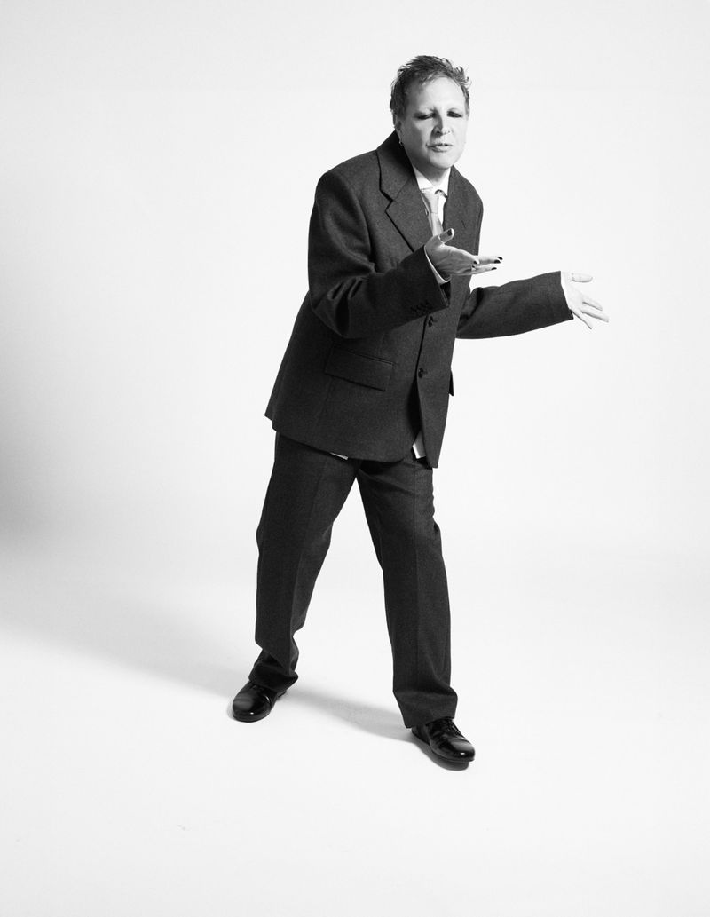 Monochrome studio portrait of a person in a suit with an open stance, hands outstretched against a stark white backdrop.