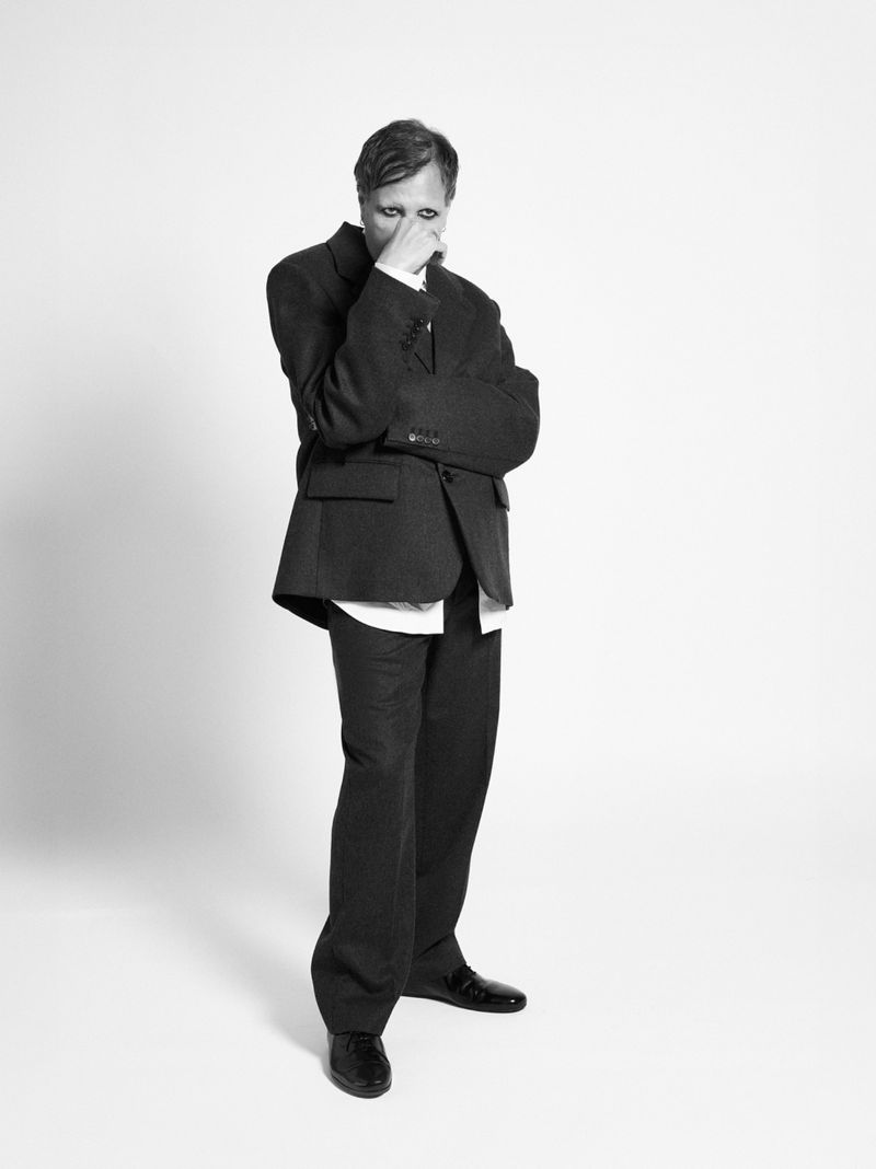 Black-and-white studio shot of a man in a suit looking down, covering his face with his hand; indoor setting, neutral background.