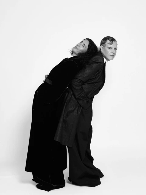 Two adult women stand back-to-back wearing black clothing in a studio shot against a white backdrop with soft, even lighting.