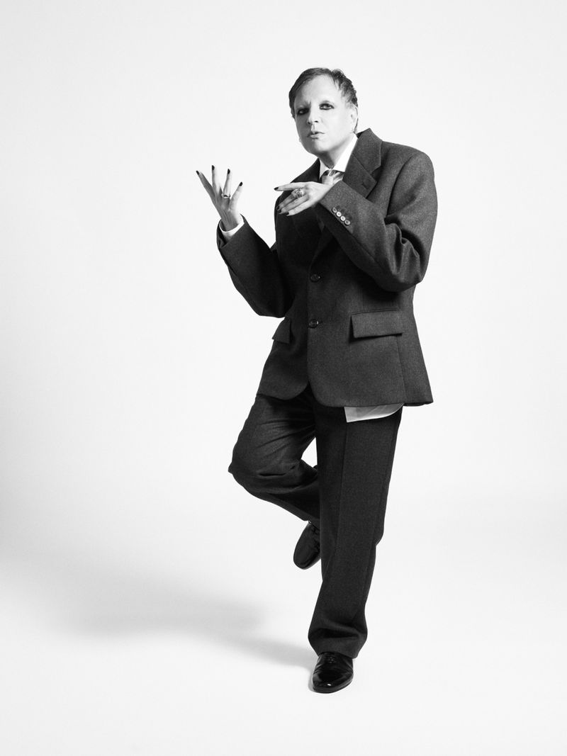 Monochrome full shot of a person in a business suit, standing with one leg raised against a plain white backdrop in a photo studio.