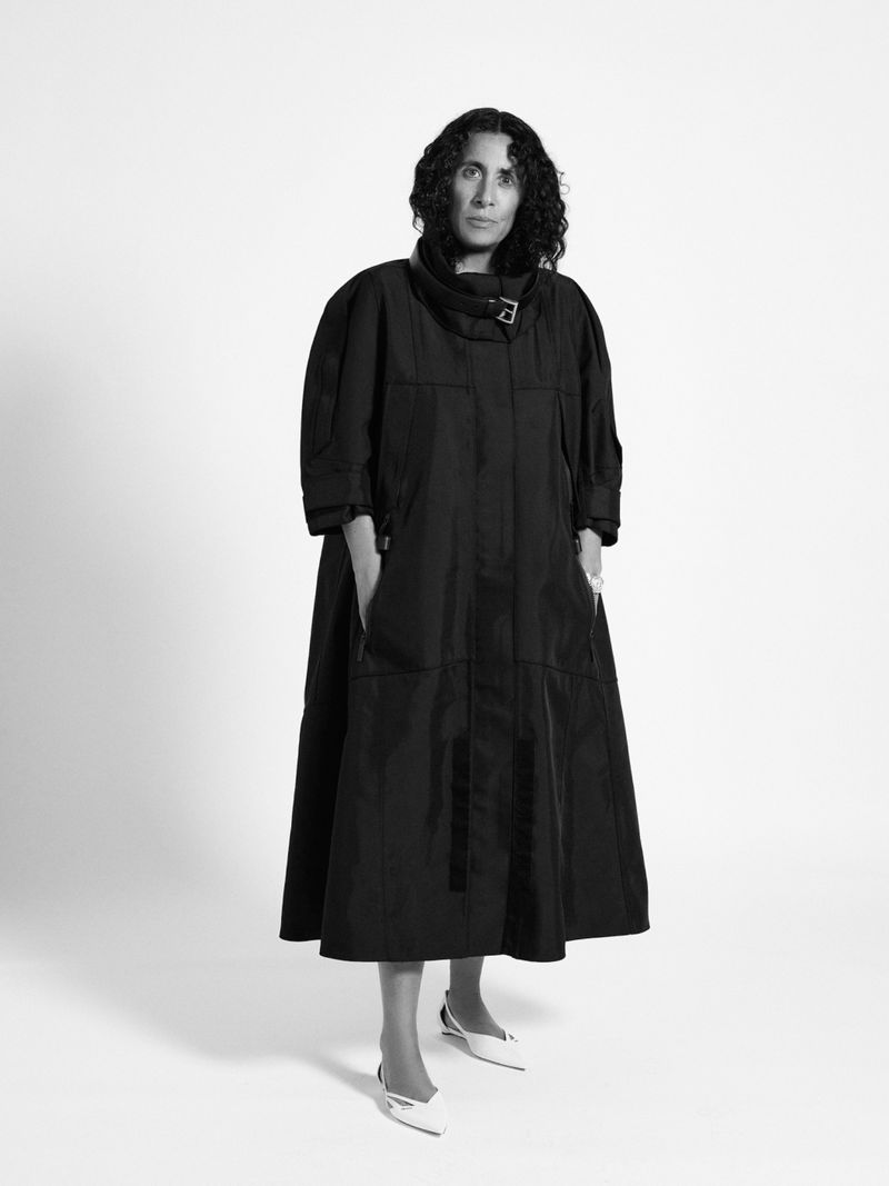 Black and white portrait of a woman with curly hair wearing a black coat and white shoes standing against a white background in a studio.