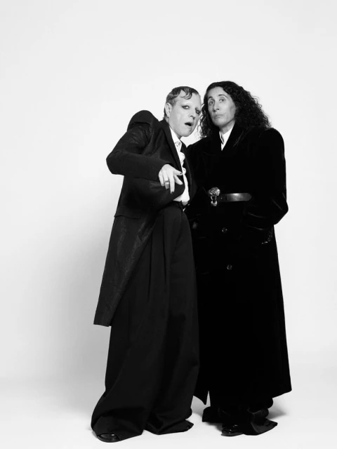 Black and white studio shot of a man with theatrical makeup in a suit with exaggerated shoulders standing next to another man in a black coat.